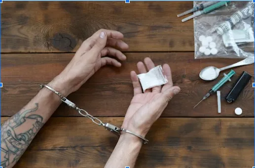 An image of a hand in handcuffs holding a sachet of white powder, with other items scattered on the table.