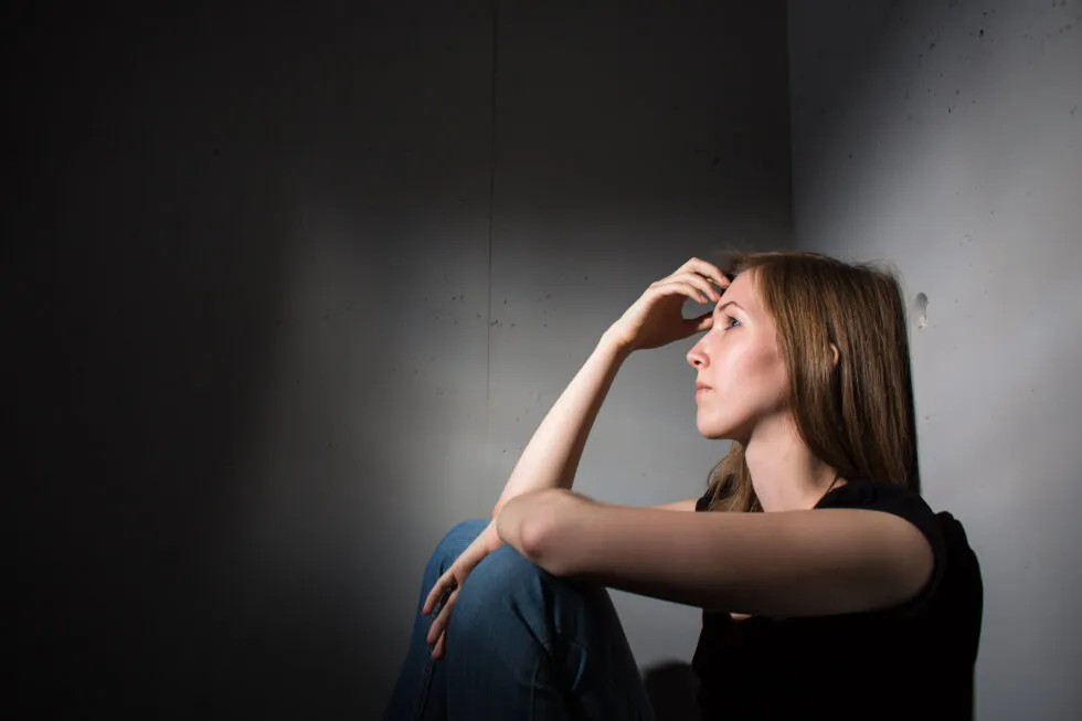 An image of a woman sitting on the floor, leaning against something, with a sad expression and a distant, lost look.