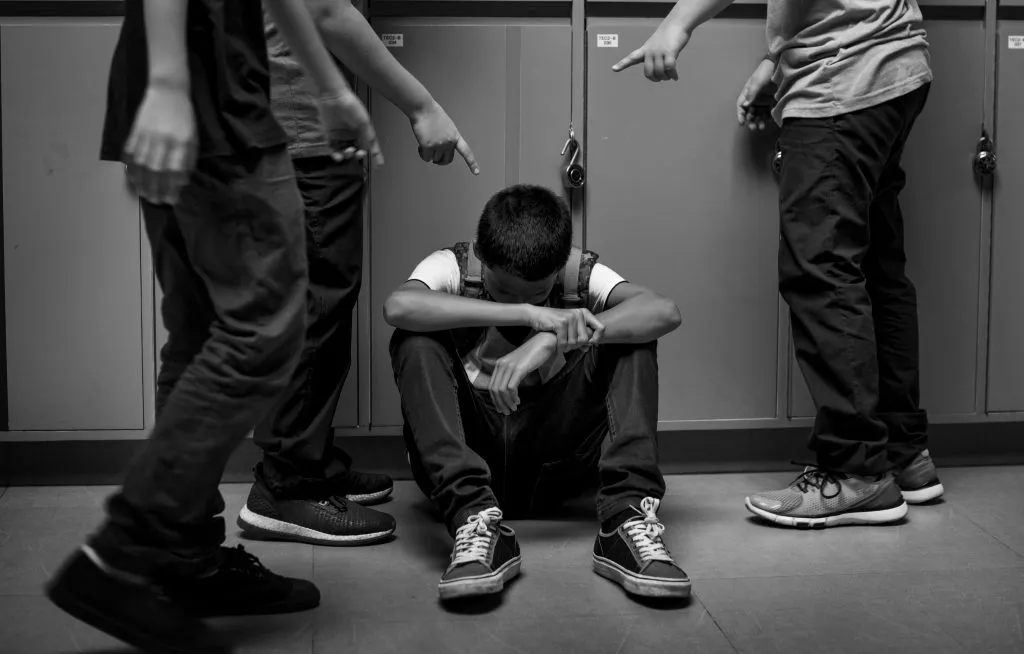 An image of a boy sitting with his head down, being bullied by other students around him.