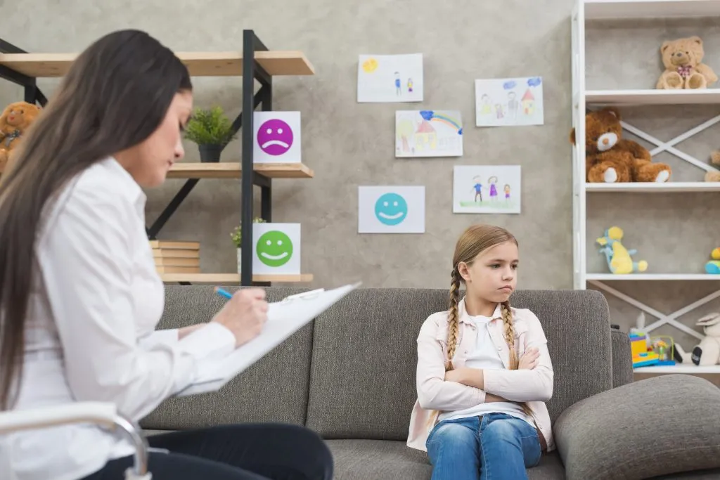 An image of a young, depressed girl sitting on a sofa, with a concerned woman sitting in front of her.