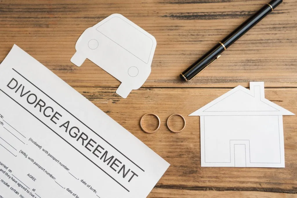 An image of a divorce agreement with a pen, a pair of wedding rings, a paper car, and a paper house placed on a table.