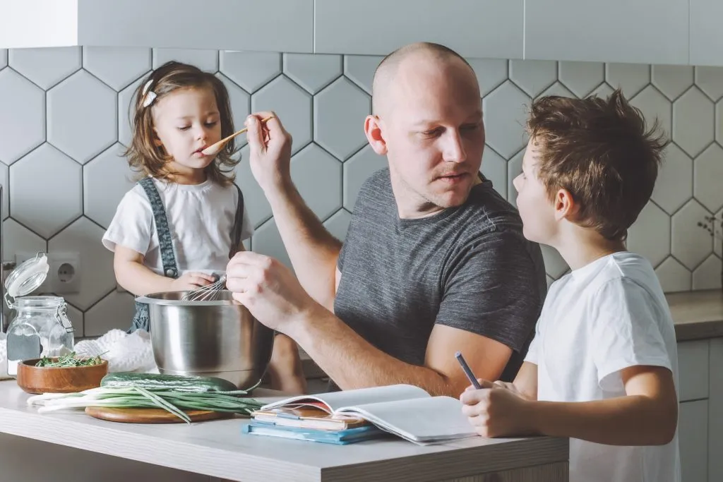An image of a father whisking an omelette in the kitchen, with his two young children watching nearby.