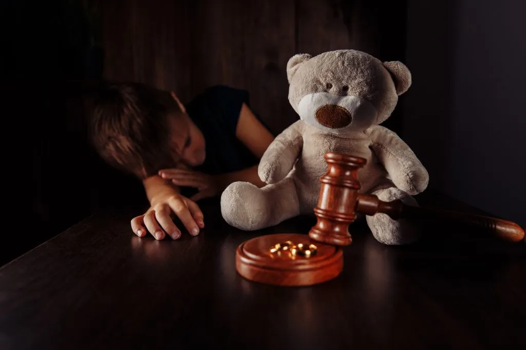 An image of a wooden gavel, a pair of rings above, and a teddy bear next to a sad young boy.