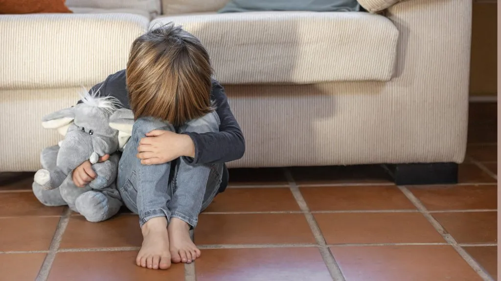 An image of a little boy sitting on the floor, upset, hugging his knees with his head bowed down.