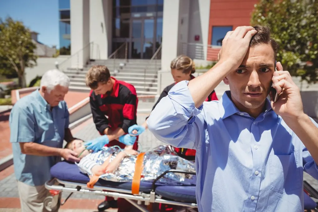 An image of a man talking on his phone, with a paramedic attending to an injured person in the background.