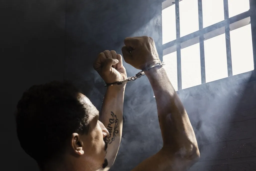 An image of a man in prison, raising his hands while wearing handcuffs