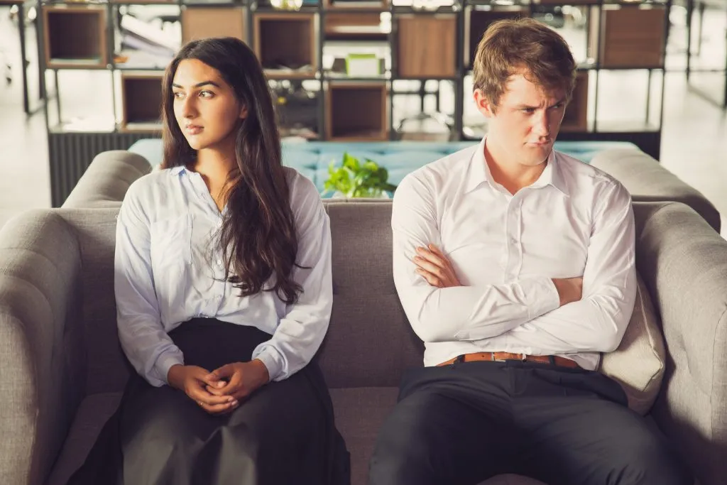 An image of a man and a woman sitting side by side on a bench, each looking in opposite directions.