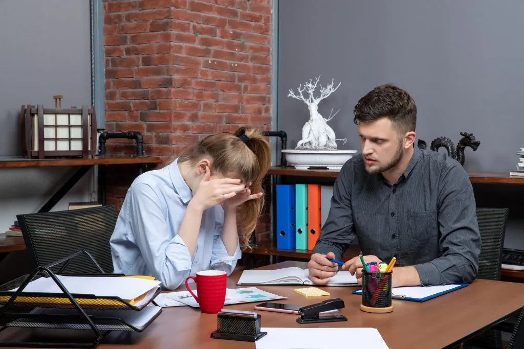 An image of a tired woman being observed by a man as she holds a pen and notebook, with various other items scattered on the table.